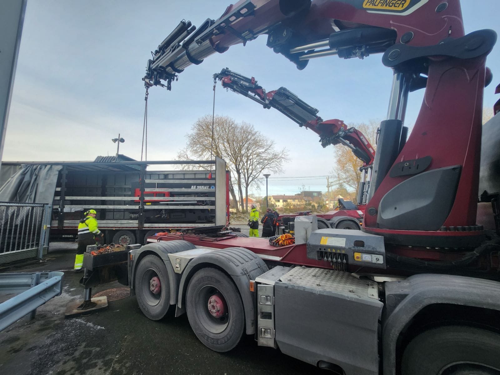 Zwei Ladekrane eines Lkw heben eine Stanzmaschine am Sattelauflieger, Monteure in Warnkleidung